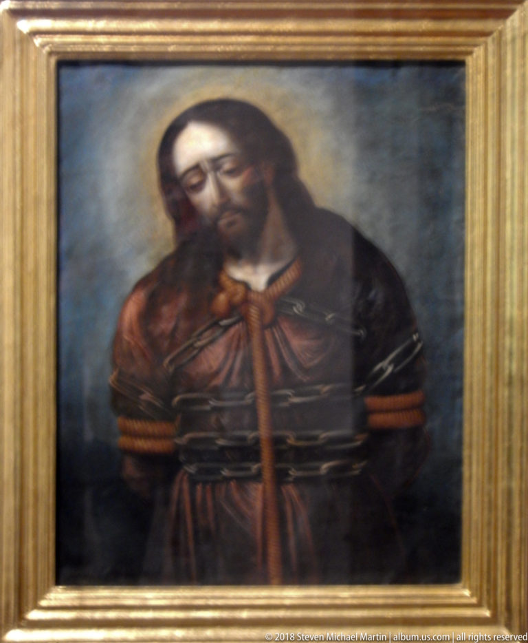 Half-length painting of Christ in a dark red robe with bowed head and hands held together at his waist.