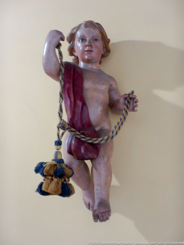 Polychrome child figure with raised arm, red drape, and blue-and-gold braided cord ending in tassels.