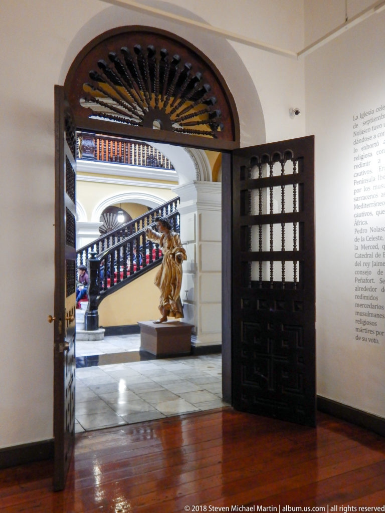 Open dark wooden doors frame a gilded figure on a pedestal with the palace staircase visible behind.