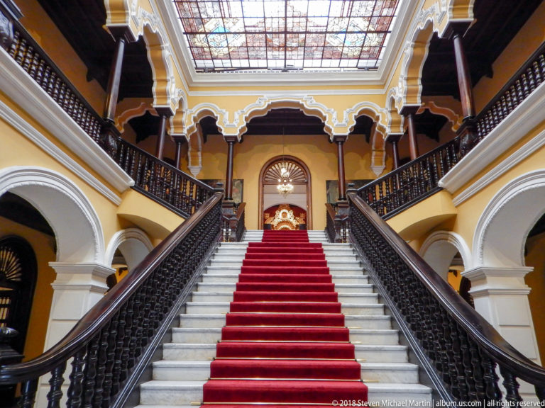 Centered white staircase with red runner and dark railings beneath a decorative glass roof and yellow upper gallery.