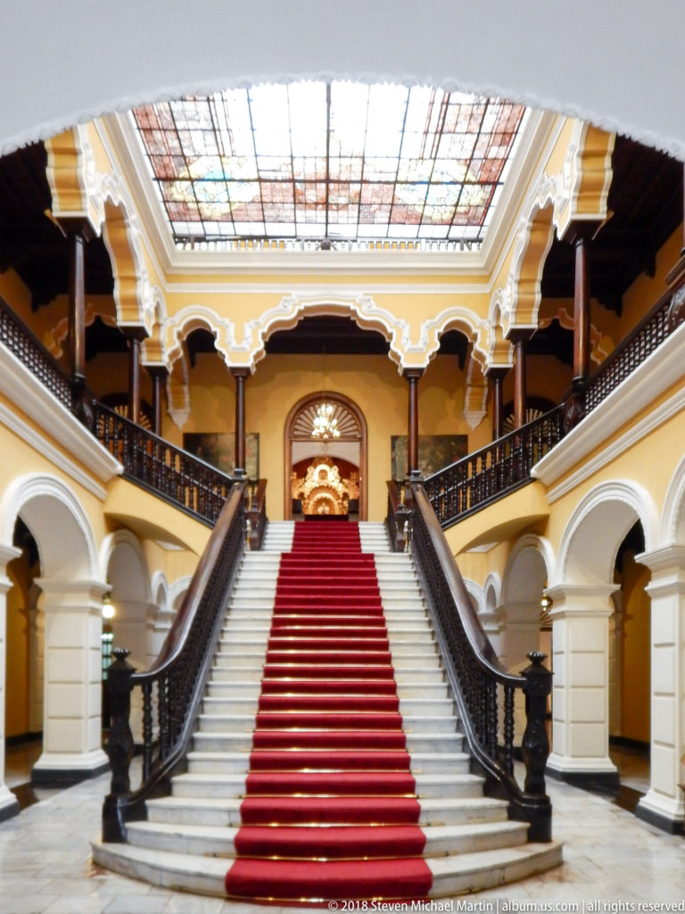 Empty white grand staircase with red runner rises between dark railings beneath yellow galleries and a stained-glass ceiling.