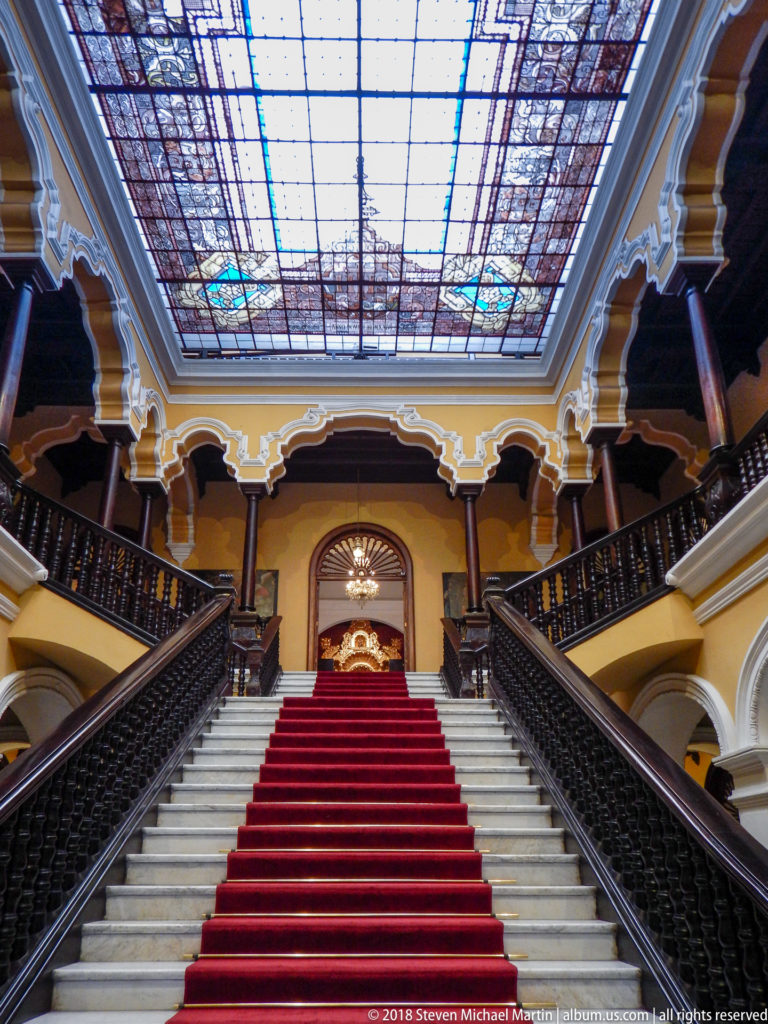 Red-carpeted grand staircase rises between dark railings beneath a large stained-glass ceiling and upper galleries.