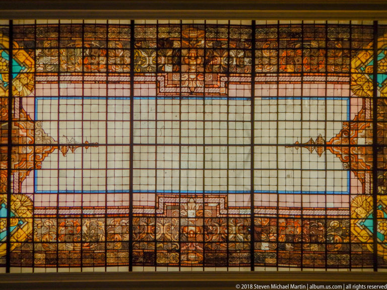 Rectangular stained-glass panel with pale central field surrounded by amber, blue, green, and red ornamental borders.