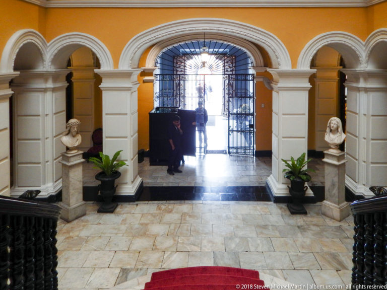 Arched entrance hall with stone floor, paired busts and plants framing an iron-gated doorway to daylight.