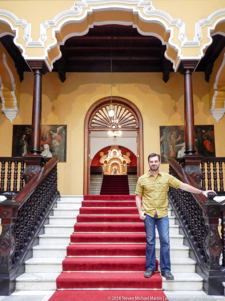 Man stands on a red-carpeted white staircase between dark carved railings, with the chapel altarpiece visible above.