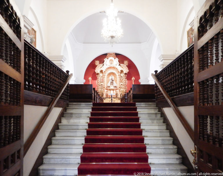 White staircase with red runner and dark wooden railings rises toward a distant gilded chapel altarpiece.