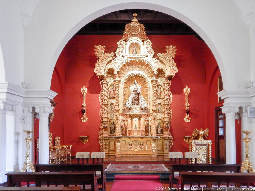 White-arched chapel with dark benches and red carpet facing a gilded retablo on a deep red sanctuary wall.