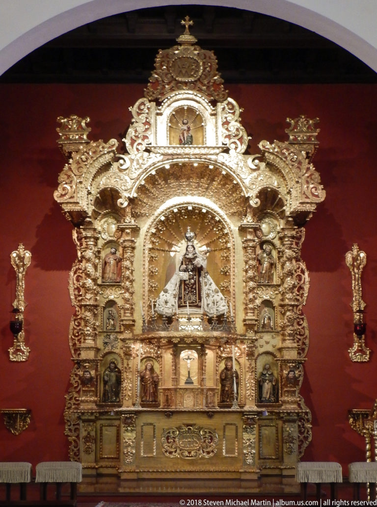 Gilded chapel retablo with central crowned Marian statue, smaller saint figures, and tall candlesticks against a red wall.
