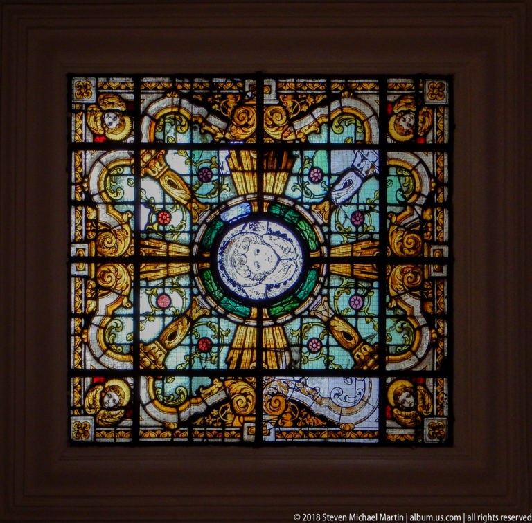 Rectangular stained-glass panel with central circular medallion, gold rays, green scrollwork, and floral ornament.