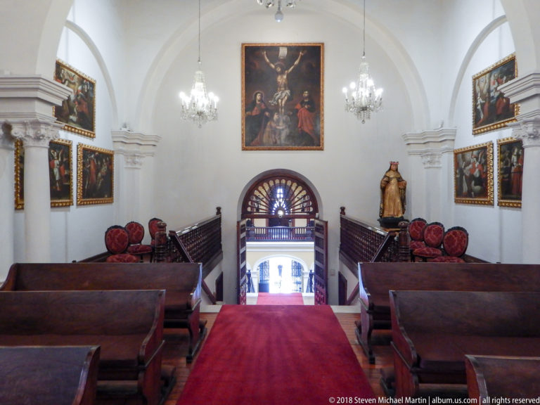 Narrow chapel with wooden benches, red carpet, chandeliers, religious paintings, and fanlight opening at the far end.