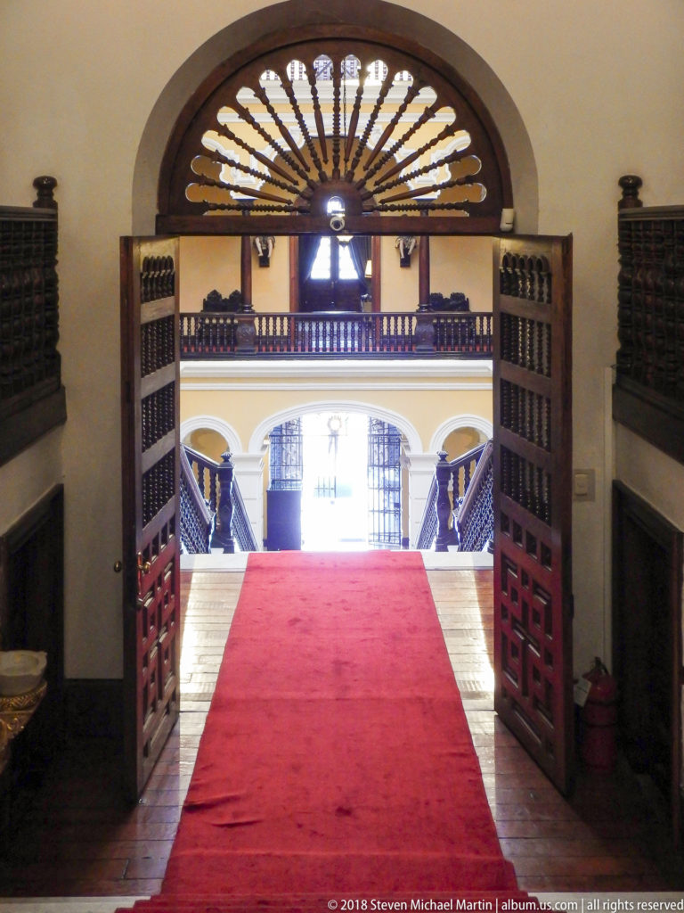 Semicircular fanlight and open wooden doors frame a red-carpeted passage toward the bright stair hall.