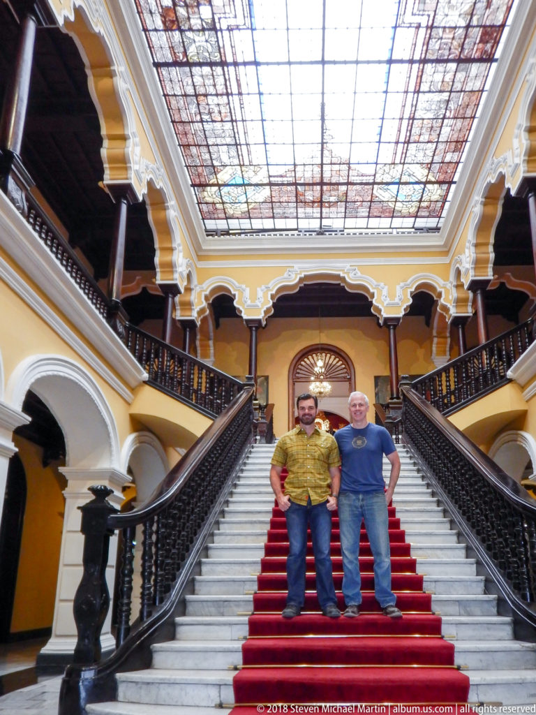 Two men stand on the red-carpeted grand staircase beneath a large decorative stained-glass ceiling and surrounding galleries.