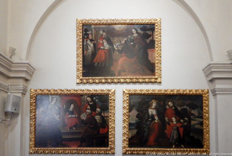 Three gilt-framed religious paintings arranged one above two on a white chapel wall.