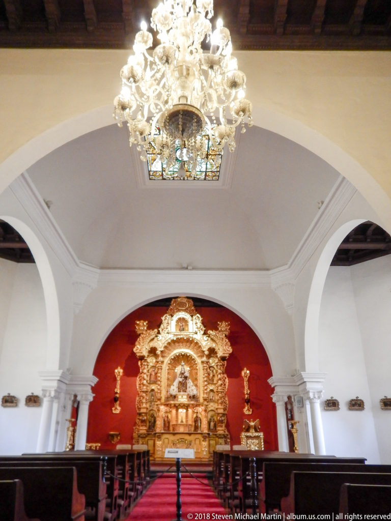 Small chapel with gilded altarpiece against red wall, white arches, chandeliers, and rows of wooden benches.