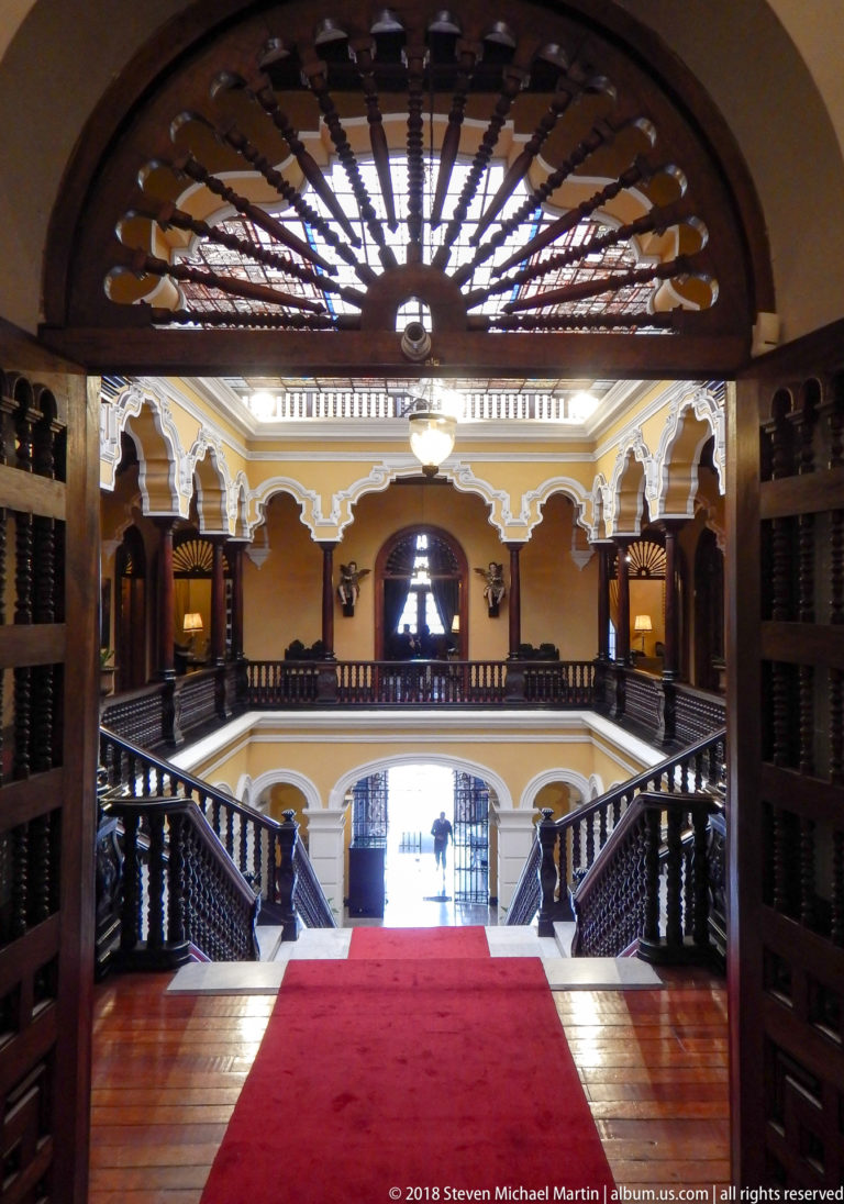 Open carved doors and fanlight frame a red-carpeted gallery overlooking the bright central stair hall.