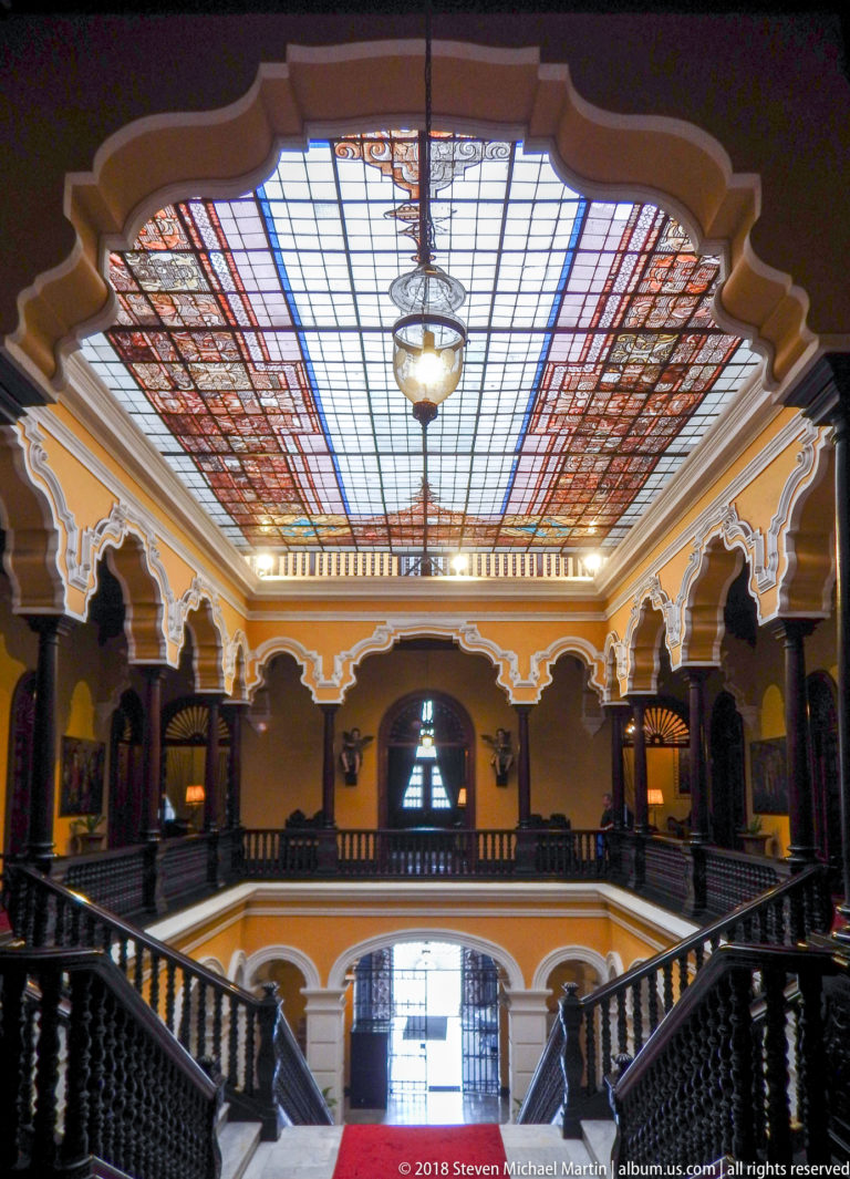 Patterned glazed ceiling with hanging lantern above white cusped arches and dark gallery railings.