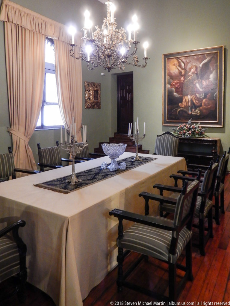 Long formal dining table with striped armchairs, candlesticks, chandelier, curtains, and large religious painting.