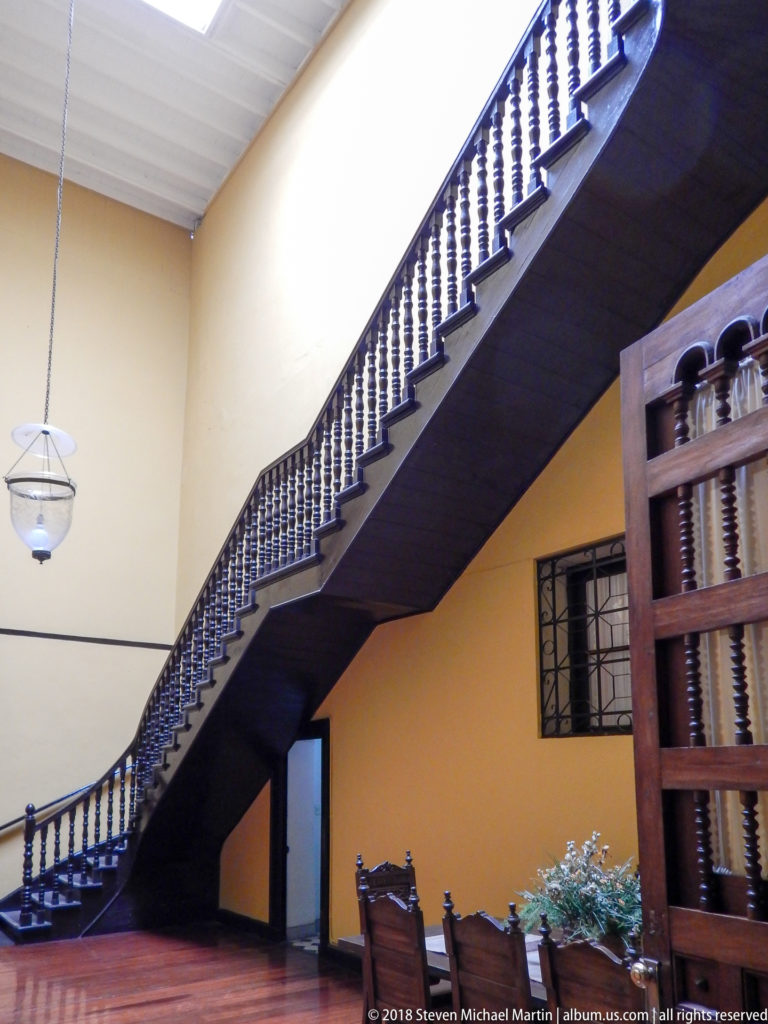 Long dark wooden staircase with turned balusters rises diagonally along a pale interior wall.
