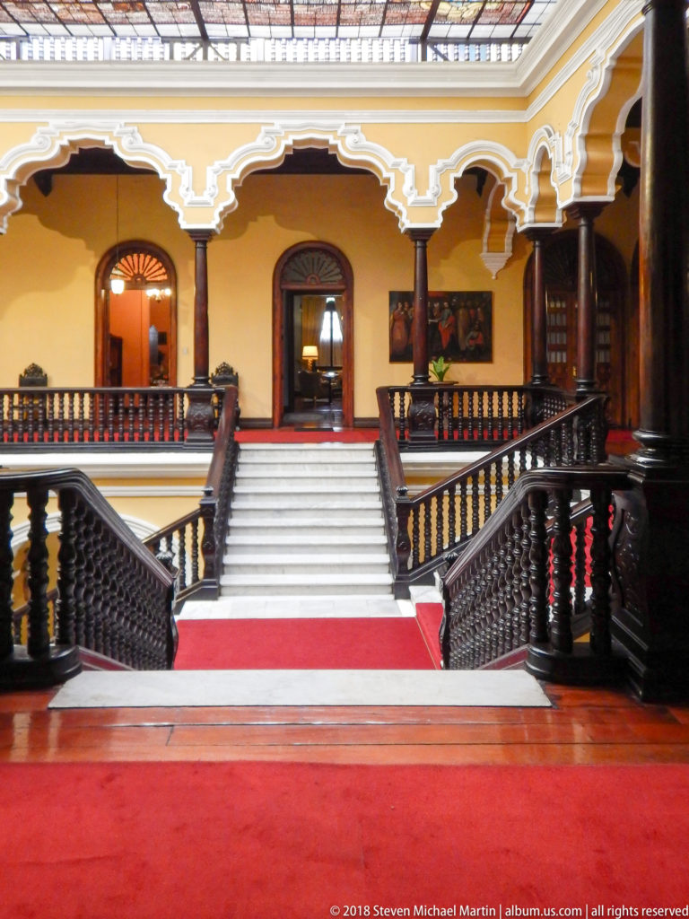 Red carpet leads to a central white staircase framed by symmetrical dark wood railings and yellow arcades.