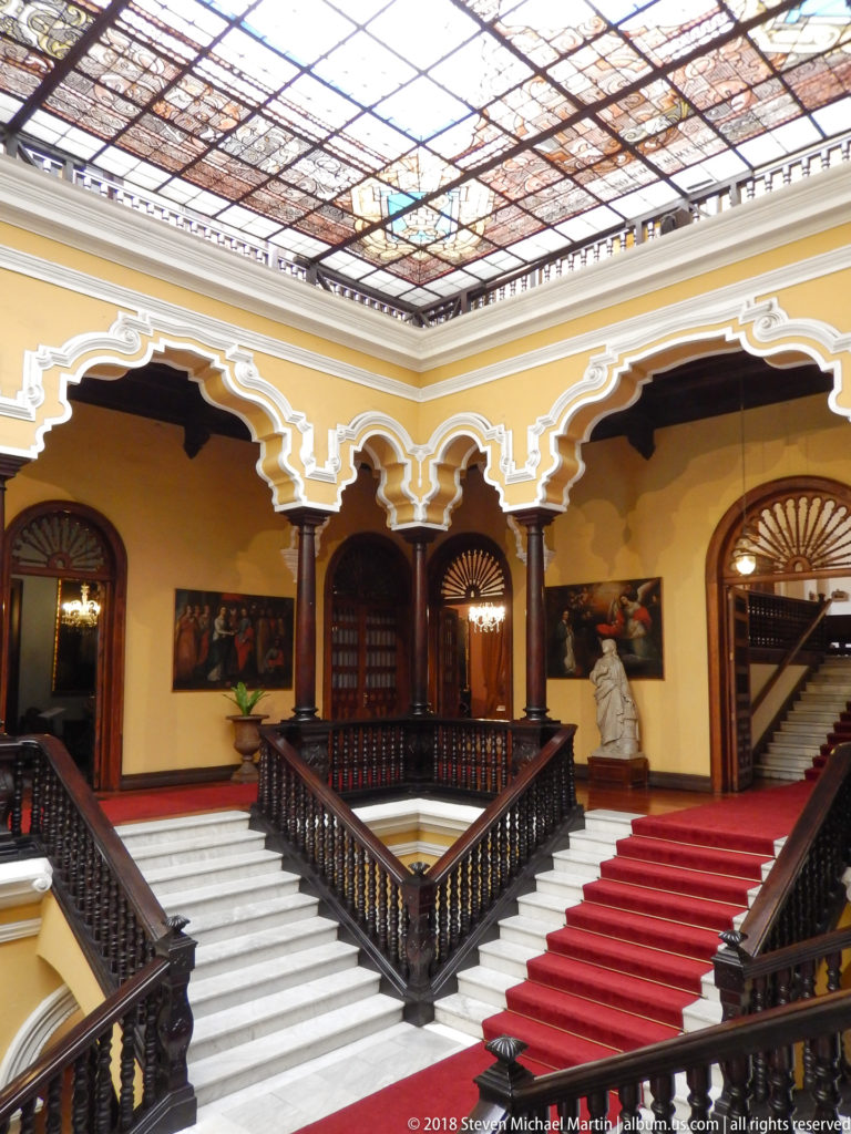 Central stair hall with paired staircases, dark balustrades, yellow walls, and patterned glazed ceiling.