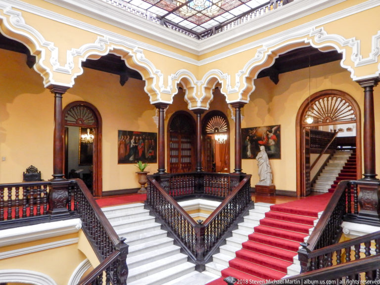 Branching white staircases with red carpet, dark balustrades, columns, and cusped arches in the central hall.