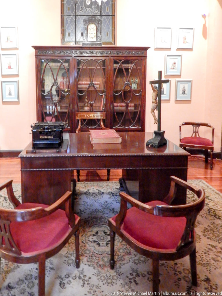 Dark writing desk with typewriter and crucifix faces red chairs before a glass-fronted bookcase.