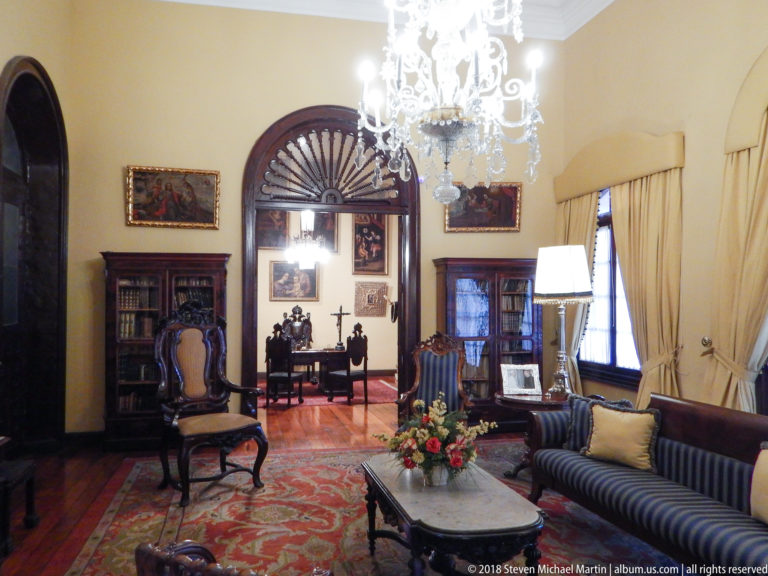 Drawing room with marble table, striped sofa, bookcases, paintings, and arched doorway to an adjoining chamber.