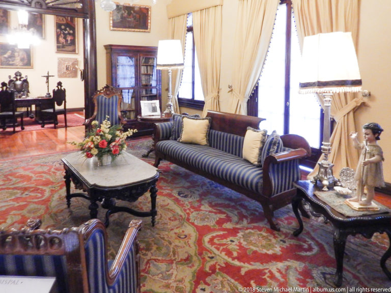 Striped sofa beside tall curtains and a marble-topped table with flowers and a small figurative sculpture.