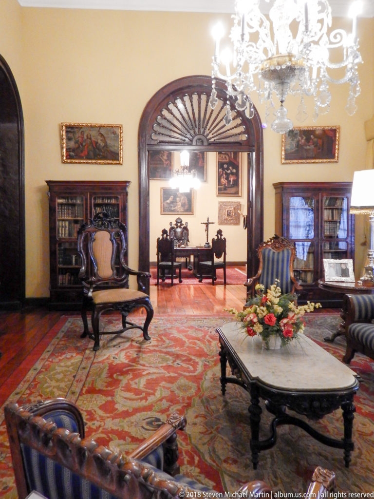 Formal drawing room with marble-topped table, carved chairs, bookcases, chandelier, and arched doorway to another room.