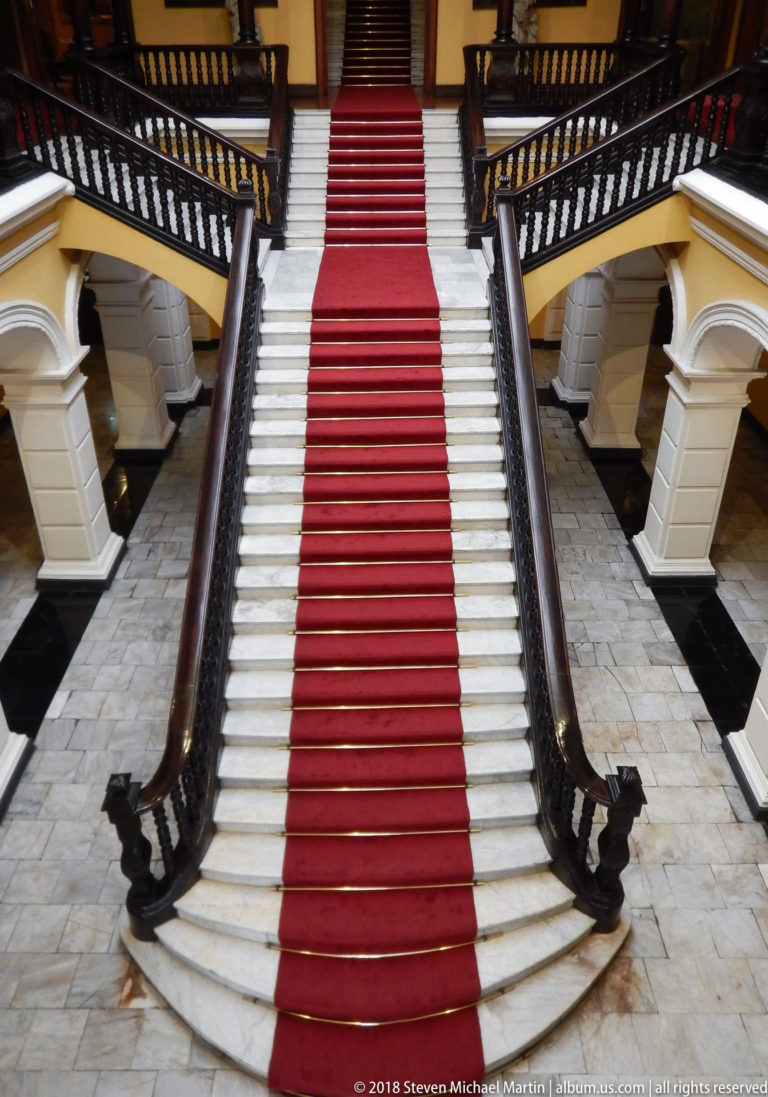 Wide white staircase with central red runner rises between dark carved wooden balustrades.