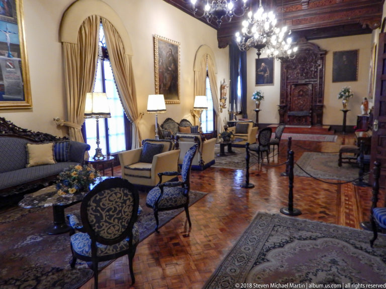 Large formal salon with patterned chairs, sofas, rugs, paintings, chandeliers, and carved wooden doorway.