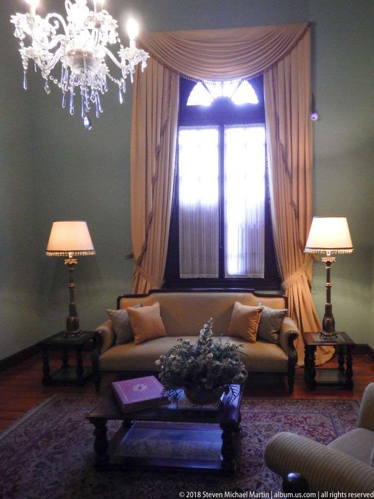 Sofa beneath a tall curtained window with matching lamps, low table, chandelier, and patterned rug.