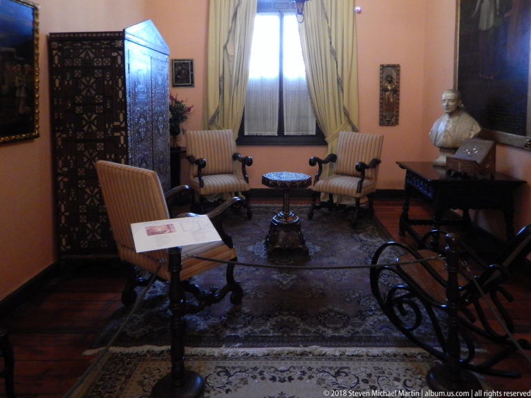 Small parlor with striped armchairs, carved screen, sculpted bust, curtained window, and patterned rug.