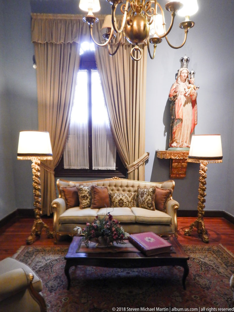 Cream sofa and matching lamps beneath a wall-mounted Virgin and Child statue in a formal sitting room.