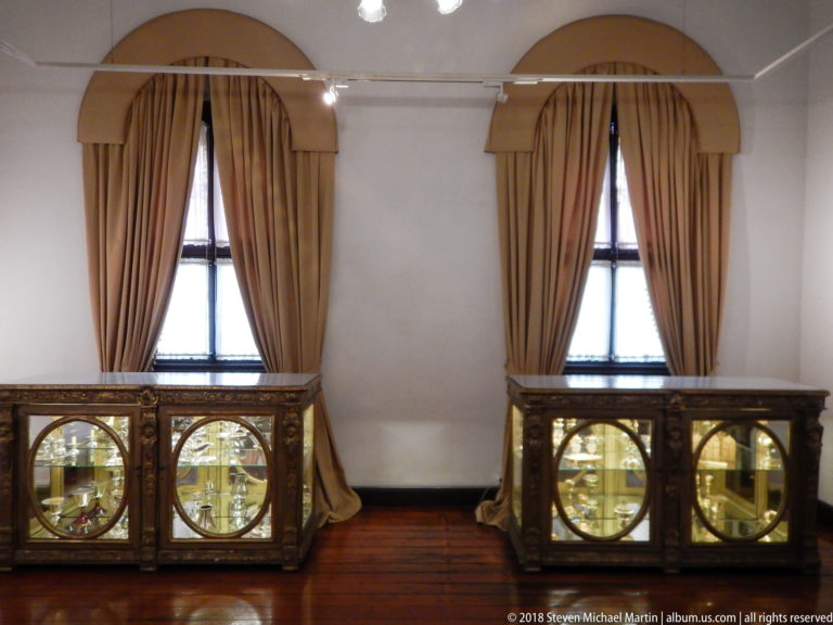 Two carved liturgical display cabinets stand beneath tall curtained windows on a polished wooden floor.
