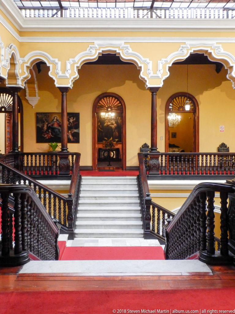 Symmetrical white staircase with red runner rises to a yellow arcaded gallery with dark wood balustrades.