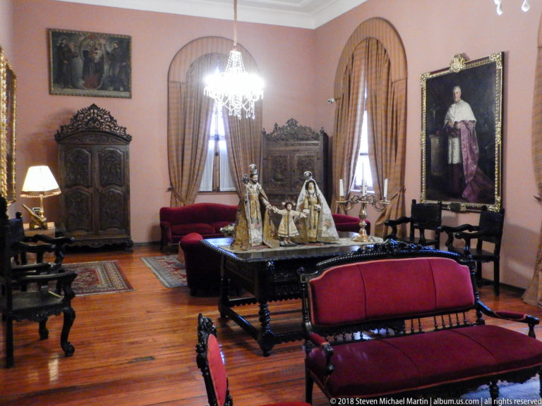Formal salon with red seating, carved cabinets, portraits, chandelier, and devotional sculpture on a central table.