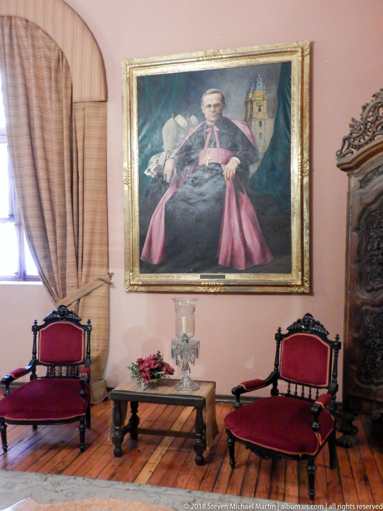 Portrait of a Catholic prelate hangs above red upholstered seating and a small table in a formal room.