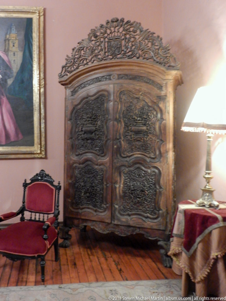 Large dark carved armoire with ornate crest stands beside red upholstered chairs in a formal room.
