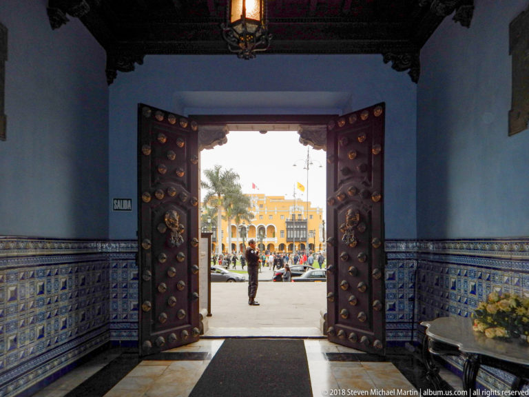 Open studded wooden doors in a tiled vestibule frame the bright Plaza Mayor outside.
