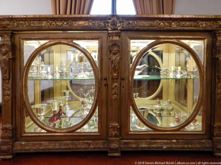 Dark carved vitrine with circular glass panels displaying numerous chalices, cups, cruets, and liturgical vessels.