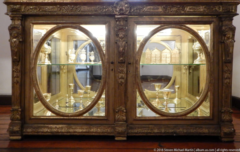 Carved dark wooden cabinet with two circular glass doors displaying chalices and other ecclesiastical metal vessels.