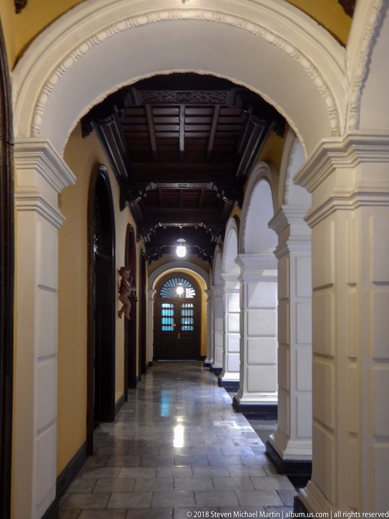 Long stone-floored corridor with white arches, yellow walls, dark timber ceiling, and closed doors at the far end.