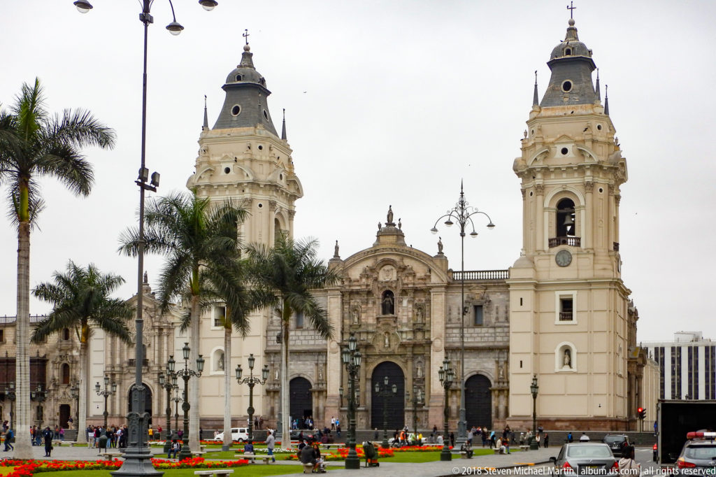 Main façade of Lima Cathedral with twin bell towers facing the Plaza Mayor, with palms, flower beds, lamps, and pedestrians.
