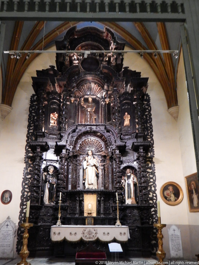 Large dark carved wooden retablo with central saint figure, smaller niche sculptures, altar table, and brass candlesticks.