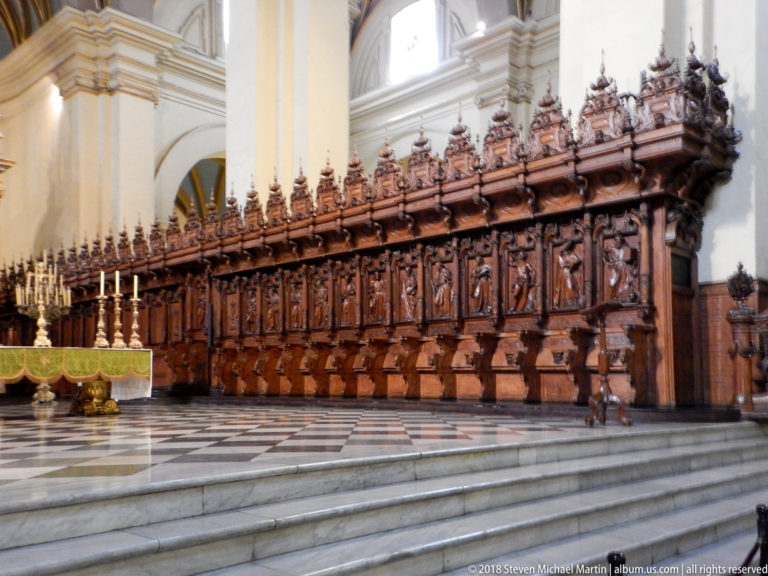 Long row of richly carved wooden choir stalls with high backs and finials, raised above marble steps beside the sanctuary.