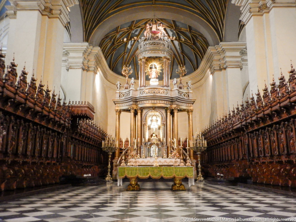 Black-and-white checkered floor leads between carved choir stalls to the high altar beneath a patterned vaulted ceiling.