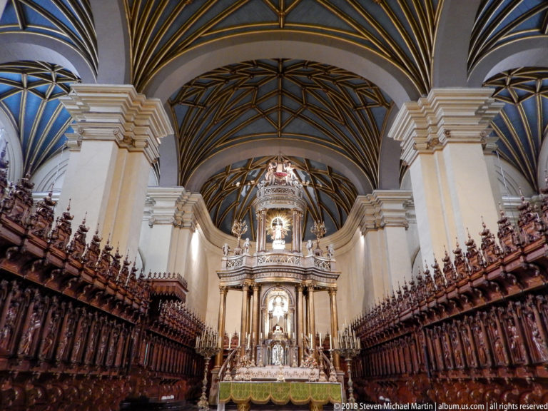 Wide symmetrical view between carved choir stalls toward the high altar beneath dark geometric vaulting and cream piers.