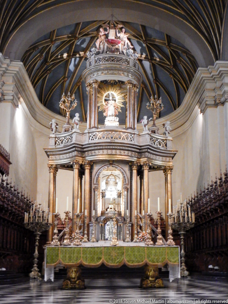 Centered sanctuary view with green-fronted high altar, monumental columned structure, carved choir stalls, and patterned vault overhead.