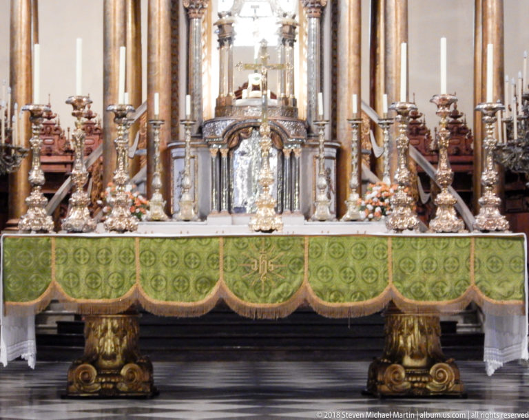 Close view of the high altar with green frontal, silver-toned candlesticks and vessels, flowers, and polished columns behind.